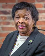 The image shows a woman posed for a professional portrait. She’s wearing a dark blazer over a light top, along with a necklace and a small circular pin on her lapel. Her hair is short and neatly styled, and she’s smiling slightly. The background appears to be a brick wall, giving the photo a warm, formal-yet-approachable feel.