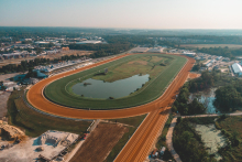 Aerial view of a horse racing track at Laurel Park, featuring a large oval dirt track surrounding a grassy infield with a pond. The surrounding area includes various buildings, roads, and greenery extending into the distance. The sky is clear, suggesting a sunny day.
